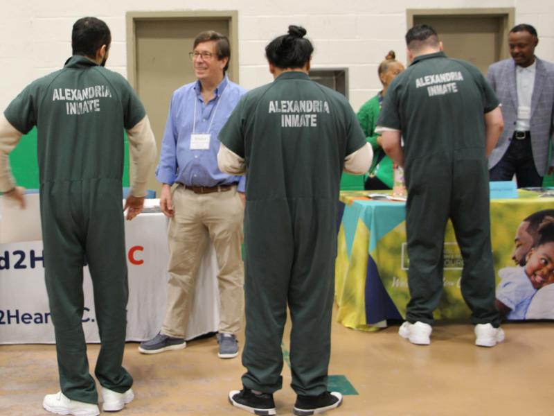Five inmates in green jumpsuits speaking with four individuals from two diferent organizations and looking at information at exhibition tables